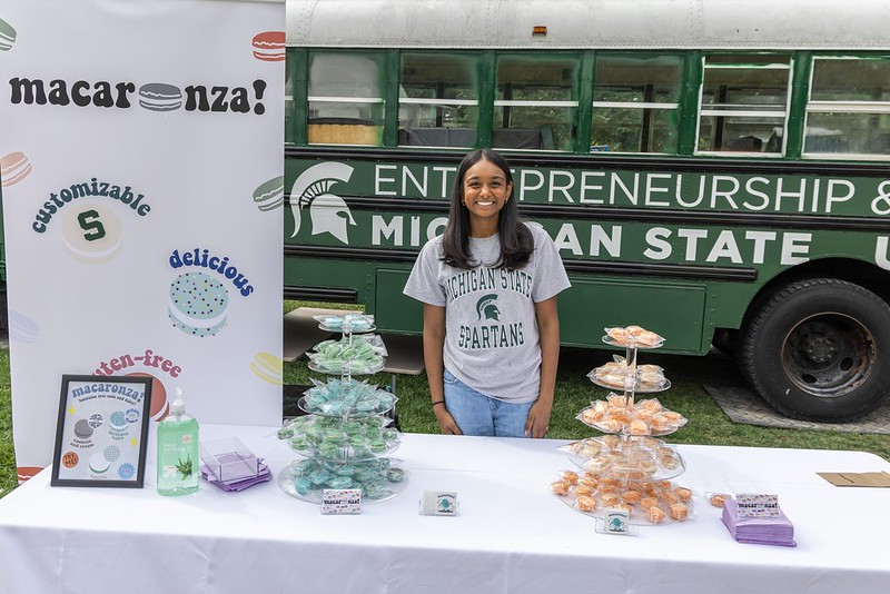 Yuktha Pulavarthi poses behind her Macaronza! sales table at Venture Summit tailgate with the MSU Startup Bus in the background.